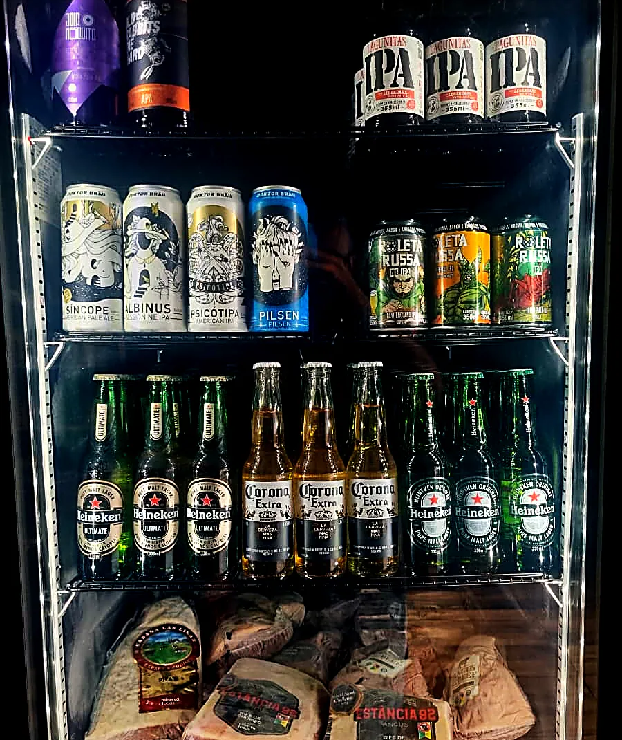Geladeira iluminada com cervejas artesanais, Heineken e Corona, e cortes embalados na prateleira de baixo